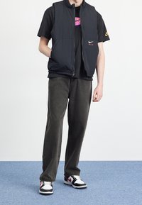 Svart ärmlös väst över en svart t-shirt med en rosa logotyp, kombinerad med mörkgröna byxor och svarta sneakers med röda och vita accenter.