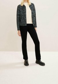 Une personne portant une veste texturée noire et blanche, un pull crème, un pantalon noir ajusté et des bottes chevilles noires  épaisses, se tenant sur un sol clair.