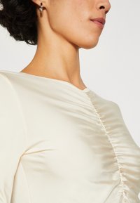 Cremefarvet satin top med bløde samlingsdetaljer langs siden. Den har en rund halsudskæring og en glat tekstur. Enkel, elegant design.