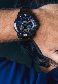 Melnā metāla rokas pulksteinis ar zilu ciparnīcu, indikatoriem, diviem sekundārajiem ciparnīcām un datuma displeju, ar izturīgu aproci un safīra kristālu.