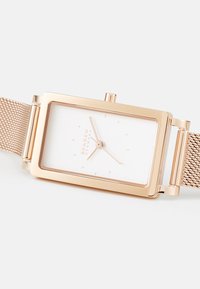Rechteckige Metalluhr mit roségoldener Beschichtung, weißem Zifferblatt, Mesh-Armband sowie minimalistischen Stundenmarkierungen und Händen. Markenname auf dem Zifferblatt.
