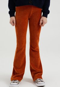 Oranžno žametne raztrgane hlače z navpičnim žametenjem, v kombinaciji z črno-belo vzorčastimi supergami. Material se zdi mehak in teksturiran.