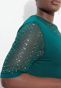 Türkise Bluse mit kurzen Ärmeln, versehen mit durchsichtiger Netztextur, die mit silbernen Punkten verziert ist. Der weiche Stoff und das strukturierte Design betonen die Silhouette.