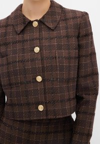 Brun kort jakke med et tekstureret ternet mønster, med gyldne spiral knapper og en klassisk krave. Detaljer i mørke tråde tilføjer dybde.