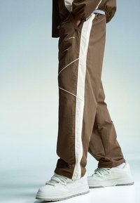 Brown track pants with white side stripes, made of lightweight fabric. Features loose fit and pockets. Paired with white sneakers.