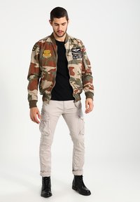 Camouflage bomberjack met patches, zwart T-shirt, cargobroek en zwarte laarzen. Gestructureerde stof, pasvorm en militaire ontwerpaccenten.