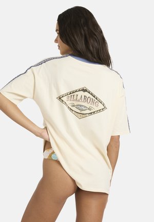 T-shirt surdimensionné couleur crème avec un logo en forme de diamant, présentant des accents bleus le long des manches et une silhouette décontractée.