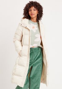 Longue doudoune beige avec capuche, présentant des panneaux matelassés, une fermeture éclair à l'avant et des fermetures à pression. Portée par-dessus un pull en maille à motifs et un pantalon vert.
