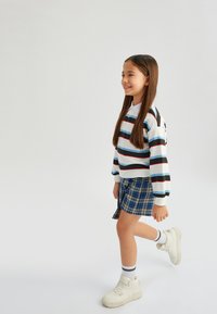 Randig långärmad topp i vitt, svart, rött och blått; mini kjol i blå rutor; vita sneakers med randiga ankelstrumpor; gångställning.