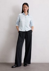Hellblaues Button-Up-Shirt mit einer einzelnen Brusttasche, kombiniert mit schwarzen, locker sitzenden Hosen und schwarzen Slippern. Glatte Stofftextur.