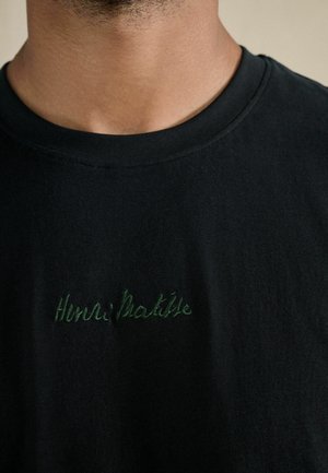Close-up of a person wearing a black crewneck shirt with "Henri Matisse" embroidered in green on the chest.
