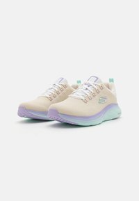 Zapatos deportivos de color beige con parte superior de malla transpirable, cordones blancos, acentos morados en la suela y etiqueta verde en el talón. Ligeros y con un diseño acolchado.