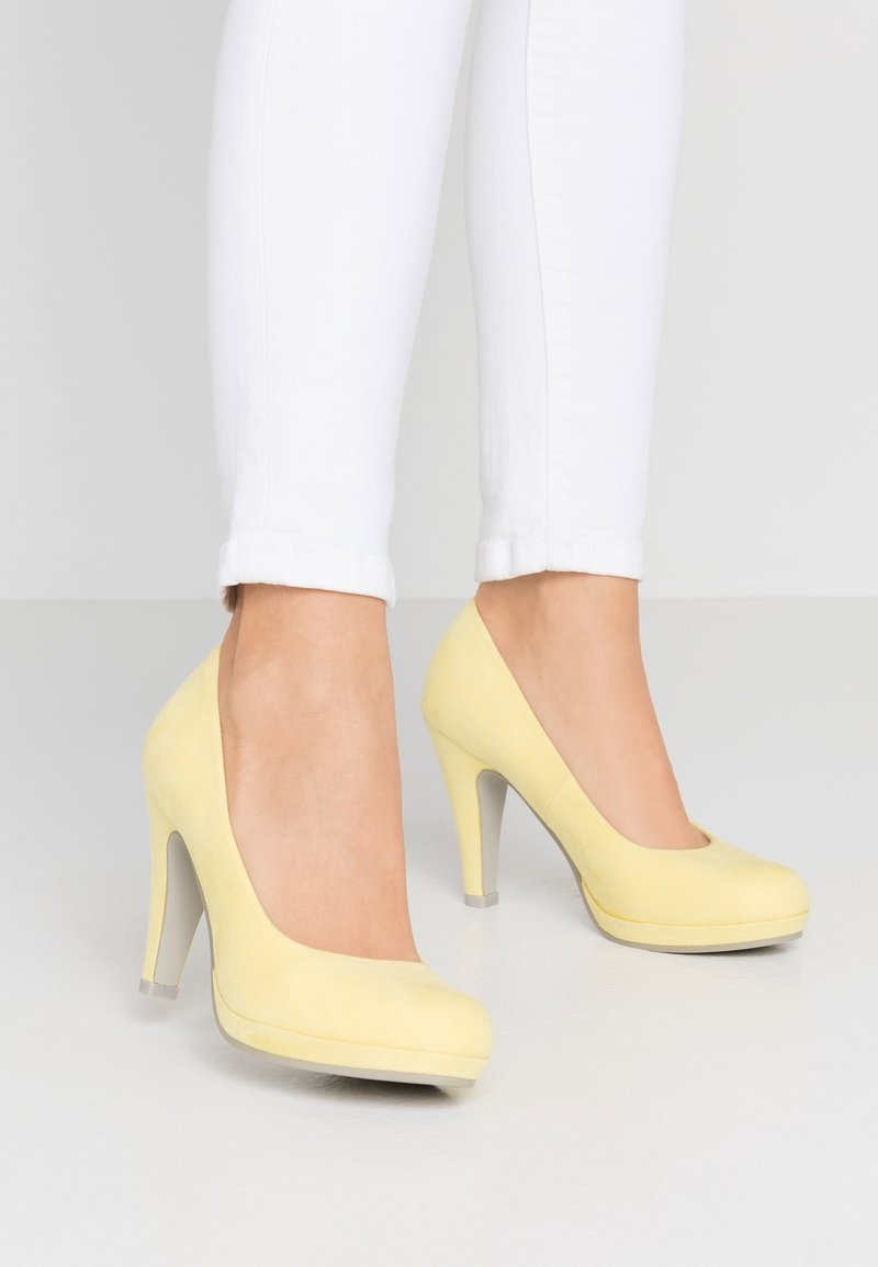 Chaussures à talons hauts en daim jaune avec un design à bout rond, présentant une texture lisse et un accent transparent sur le talon. Portées avec un jean blanc retroussé.