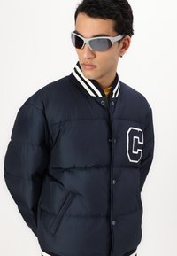 Námornícka pufrová bunda s bielym pruhovaným golierom, zdobená logom 'C' na ľavej strane a dvoma postrannými vreckami. Hladká, polstrovaná textúra.