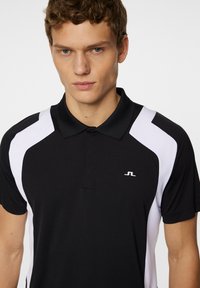 Svart pikétröja med vita detaljer på axlarna, med klassisk krage och korta ärmar. Inkluderar en liten logotyp på bröstet.