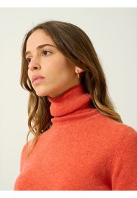 Turtleneck-Pullover aus leuchtend orangefarbenem Strickstoff. Hat einen eng anliegenden Kragen und eine glatte Textur. Kleine Creole betont den Look.