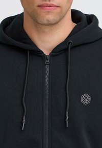 Svart hettegenser med glidelås laget av mykt stoff, med snører, glidelås foran og en liten brodert logo på brystet.