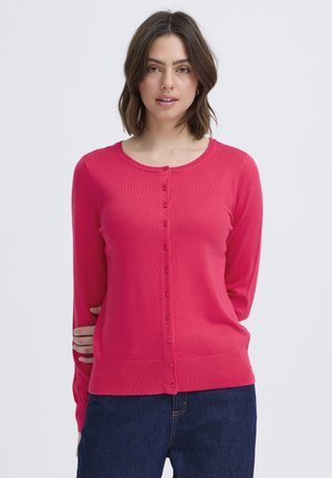 Donna con capelli castani fino alle spalle indossa un cardigan rosa brillante con bottoni e jeans blu scuro su uno sfondo chiaro e uniforme.