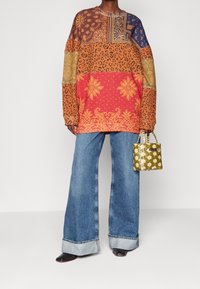 Blusa de manga larga con estampado que presenta tonos naranjas, azules y dorados, combinada con vaqueros de mezclilla acampanados y un pequeño bolso verde con textura de panal.