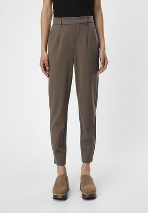 Femme portant un pantalon marron taille haute fuselé avec passants de ceinture et des sabots à plateforme taupe avec bordure cloutée, debout devant un fond uni.