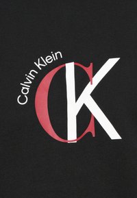 Czarna koszulka z dużym czerwono-białym logo "CK" oraz tekstem "Calvin Klein" w zakrzywionym kroju nad logo. Miękka tekstura tkaniny.