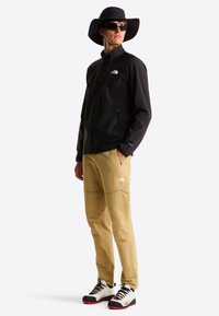 Μαύρο μπουφάν με φερμουάρ, μπεζ cargo παντελόνια με λογότυπο, καπέλο με φαρδύ γείσο και μαύρα γυαλιά ηλίου· εξοπλισμός για υπαίθριες δραστηριότητες με ανθεκτικά υλικά και αντιθετικά χρώματα.