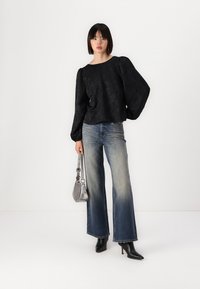 Blusa nera con texture e maniche a sbuffo, abbinata a jeans a zampa d'elefante azzurri chiari. Gli accessori includono una borsa argentata e stivali neri con tacco alto.