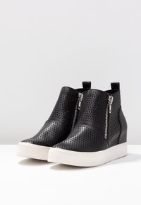 Botas de tobillo negras perforadas con cremalleras plateadas, suelas de goma blanca y una textura suave. El diseño incluye un corte alto con un leve cuña.