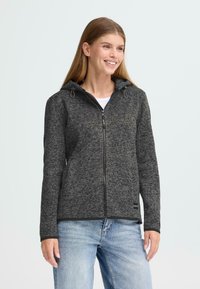 Grijze gebreide jas met capuchon, voorzien van een volledige rits, zwarte afwerking en zijzakken. Draag je over een wit shirt en blauwe jeans.