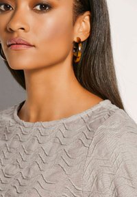 Grauer, strukturierten Pullover mit welligem Muster; große Ohrringe aus Schildpatt. Glattes, glattes Haar, das teilweise die Schulter bedeckt. Weiche Beleuchtung.