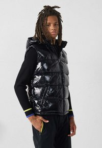 Smanicato imbottito nero con finitura lucida, cappuccio e cerniera frontale. Presenta due tasche laterali e un design aderente, indossato sopra una maglia nera a maniche lunghe.