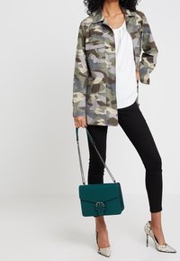 Tarnmusterjacke in Grün und Beige, kombiniert mit einem weißen Oberteil, schwarzen Skinny-Jeans und spitzen Schlangenleder-Absätzen. Gr�üne strukturierte Handtasche.