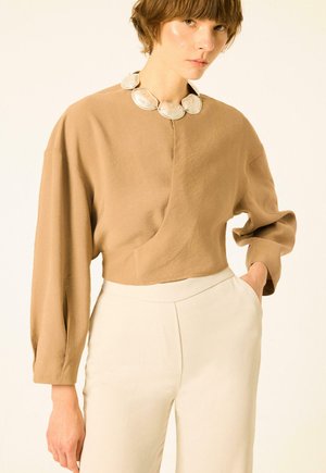 Personne portant un haut beige ample à manches longues bouffantes, un pantalon crème taille haute et un collier épais en argent, posant avec une main dans la poche.