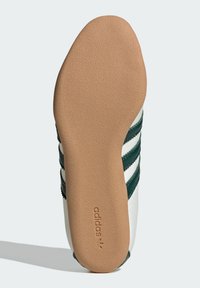 Adidasova cipela s teksturiranim smešnim gumom na dnu. Ima belu gornju stranu s zelenim prugama i logotipom. Glatka površina i zaobljeni potplat.
