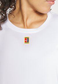 Camiseta deportiva blanca con un pequeño parche del logo de Nike, colorido, en el pecho. La tela parece suave y tiene un escote redondo.