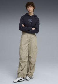 Ung person med kort, krøllet hår iført en mørk hættetrøje, beige cargo-bukser og hvide Puma sneakers, stående med armene korslagt.