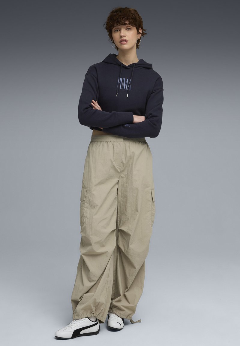 Ung person med kort, krøllet hår iført en mørk hættetrøje, beige cargo-bukser og hvide Puma sneakers, stående med armene korslagt.