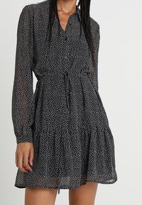 Abito nero a pois con maniche lunghe, chiusura a bottoni sul davanti, vita aderente e orlo arricciato. Realizzato in un tessuto leggero e trasparente.