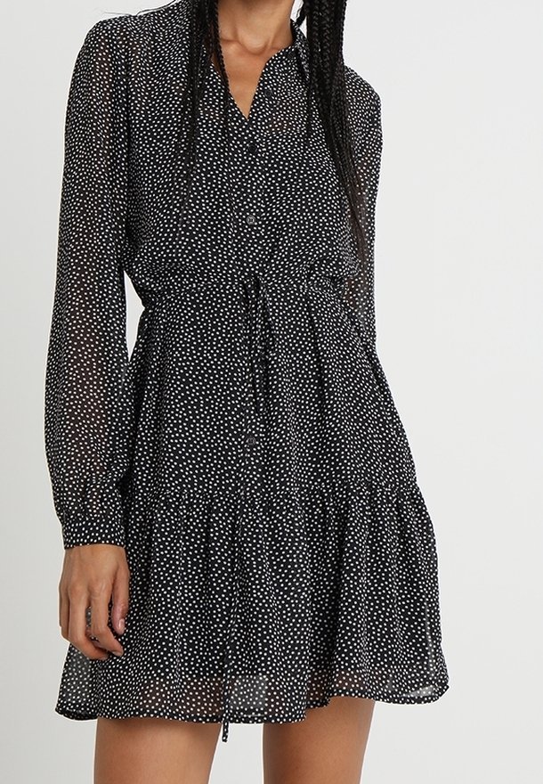 Abito nero a pois con maniche lunghe, chiusura a bottoni sul davanti, vita aderente e orlo arricciato. Realizzato in un tessuto leggero e trasparente.
