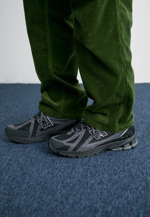 Black and gray athletic shoes with mesh panels and textured soles, paired with green corduroy pants on a blue carpet.