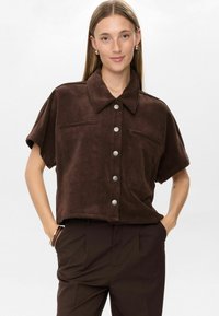 Young woman with straight hair wears a loose, short-sleeve brown corduroy button-up shirt and dark brown pants with hands in pockets.