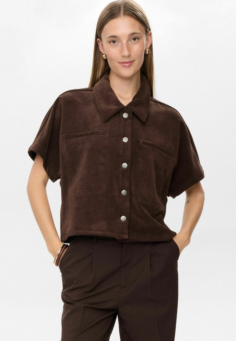 Young woman with straight hair wears a loose, short-sleeve brown corduroy button-up shirt and dark brown pants with hands in pockets.