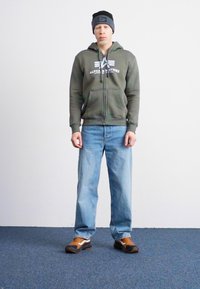 Hanorac cu fermoar de culoare verde măslin cu logo, blugi de culoare bleu deschis și adidași căprui. Căciulă neagră pe o persoană care stă în fața unui fundal simplu.