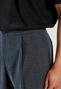 Grå plisserede bukser lavet af struktureret stof med knaplukning og sidelommer, med en glat finish sammen med en sort skjorte.