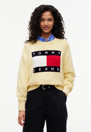 Femme aux cheveux bouclés portant un pull jaune avec un logo Tommy Jeans rouge, blanc et noir et un pantalon noir avec une ceinture.