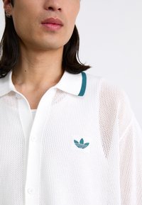 Hvit polo med nettingmateriale og en tealfarget krage. Har en liten tealfarget logo på venstre side. Teksturert stoff med knappestolpe.