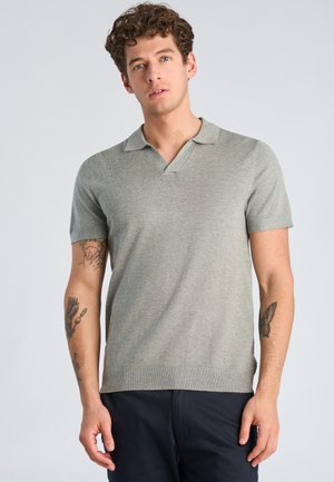 Junger Mann mit lockigem Haar, der ein hellgraues, kurzärmeliges Poloshirt und dunkle Hosen trägt und Tätowierungen auf beiden Unterarmen zeigt.