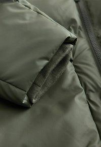 Padded alyvuogių žalia striukės rankovė, pasižyminti lygiu išoriniu paviršiumi ir minkštu mezginiu vidinėje apykaklėje. Užtrauktukas matomas palei siūlę.