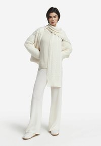 Hvidt strikket sæt bestående af en kabelstrikket sweater, bukser med brede ben og et matchende tørklæde. Blød tekstur og afslappet pasform med et hyggeligt udseende.