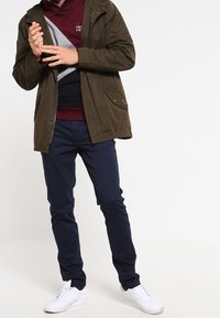 Olivgrön isolerad jacka med huva, lager över en grå och vinröd sweatshirt. Marinblå byxor och vita sneakers fulländar outfiten.